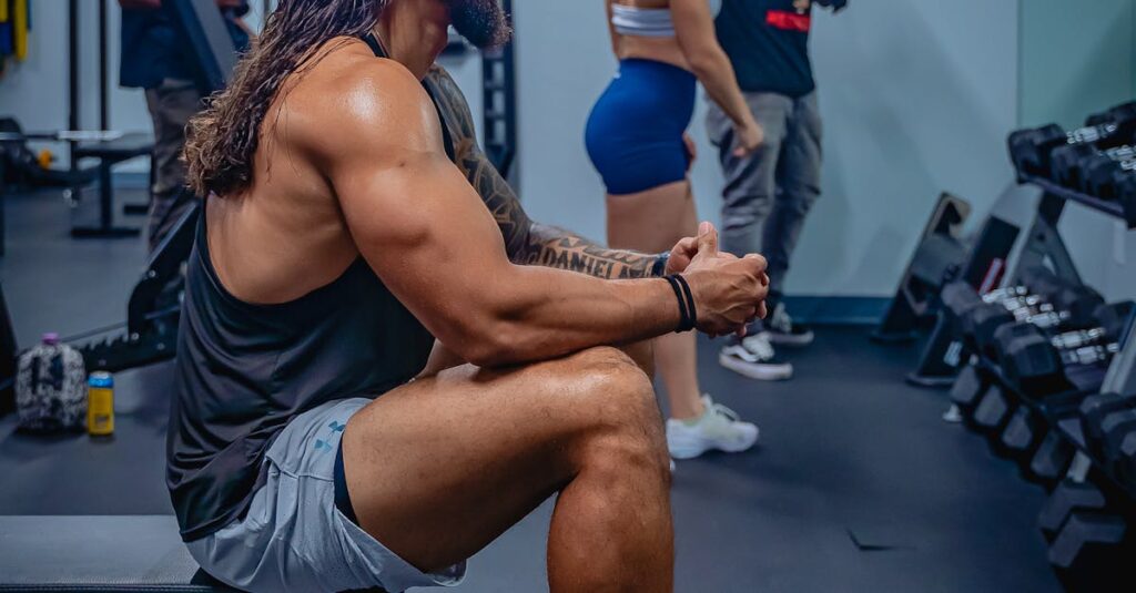 Muscular man sitting on a weight bench in a modern gym, surrounded by fitness enthusiasts.